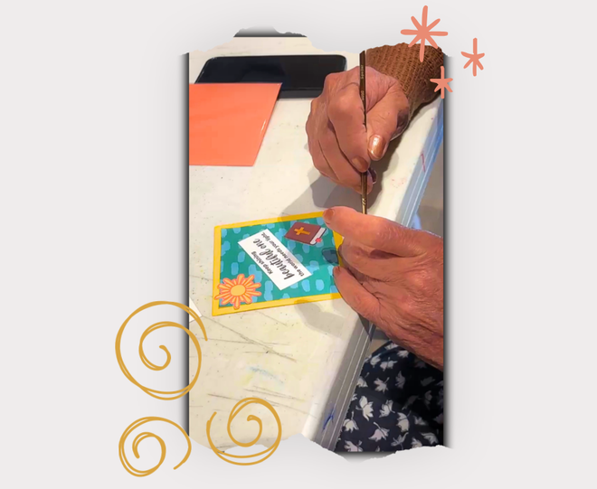A photo featuring a close-up of hands painting on a tabletop, with a small colorful card in the foreground. The print is shown against a light gray background with decorative gold swirls and pink starburst graphics.